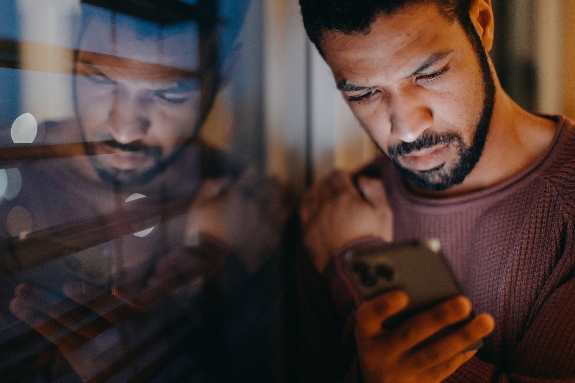 Young man with a smart phone looks sad, standing by window in evening at home